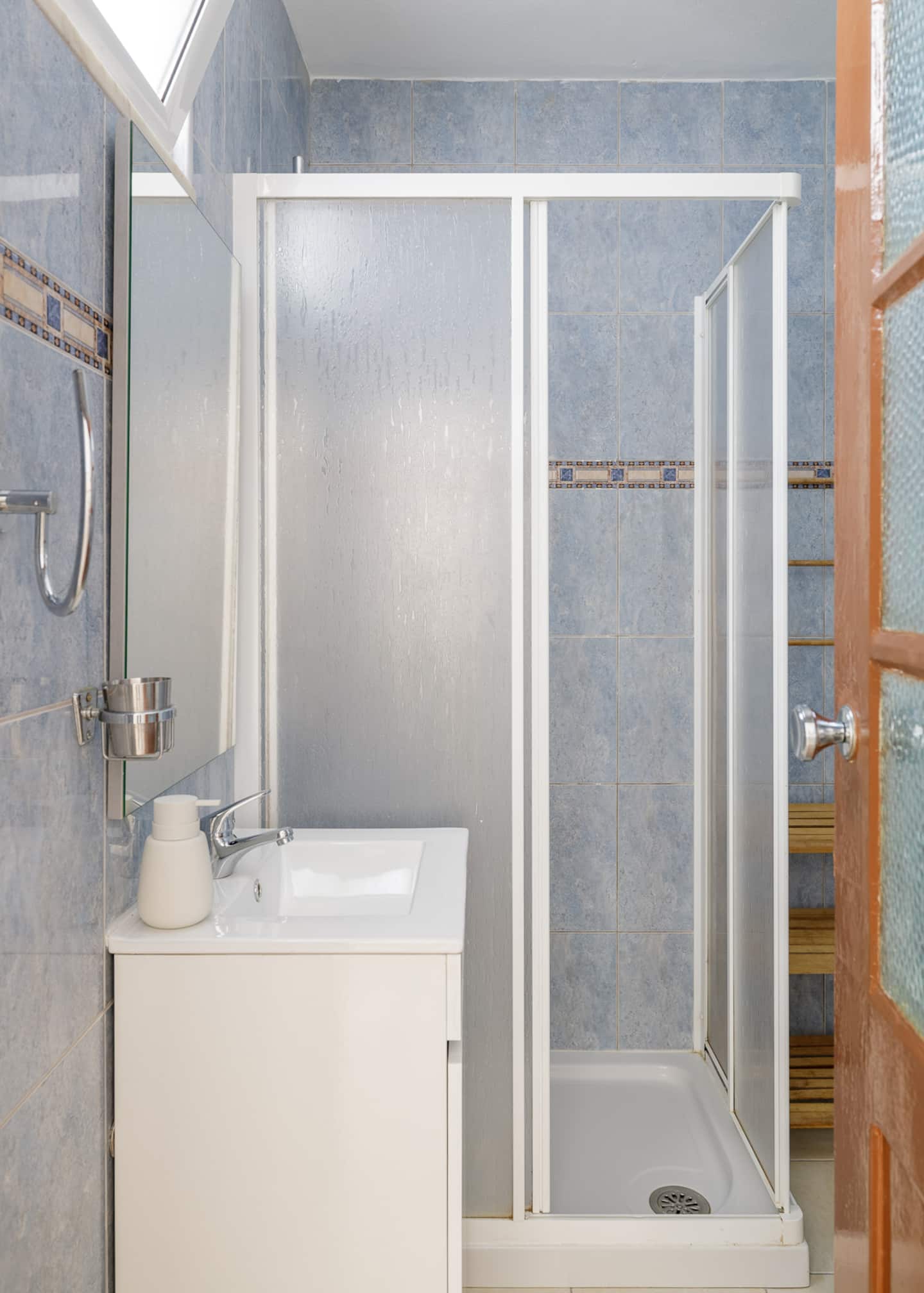 A bathroom tiled in pale blue with a patterned border: a walk-in shower cubicle with frosted glass, a white basin on a white cabinet with a mirror above, a wooden towel ladder, and a door of red timber with glass panes.