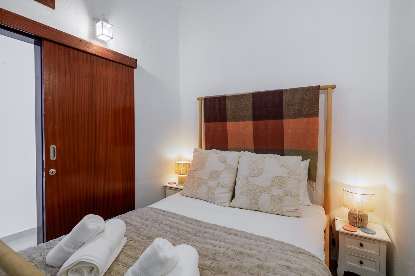 A double bed with white linen and a beige throw, towels folded on it, a headboard hung with a striped blanket in rust, brown and green, bedside lamps lit on both sides, and a sliding door of dark red timber on the left.