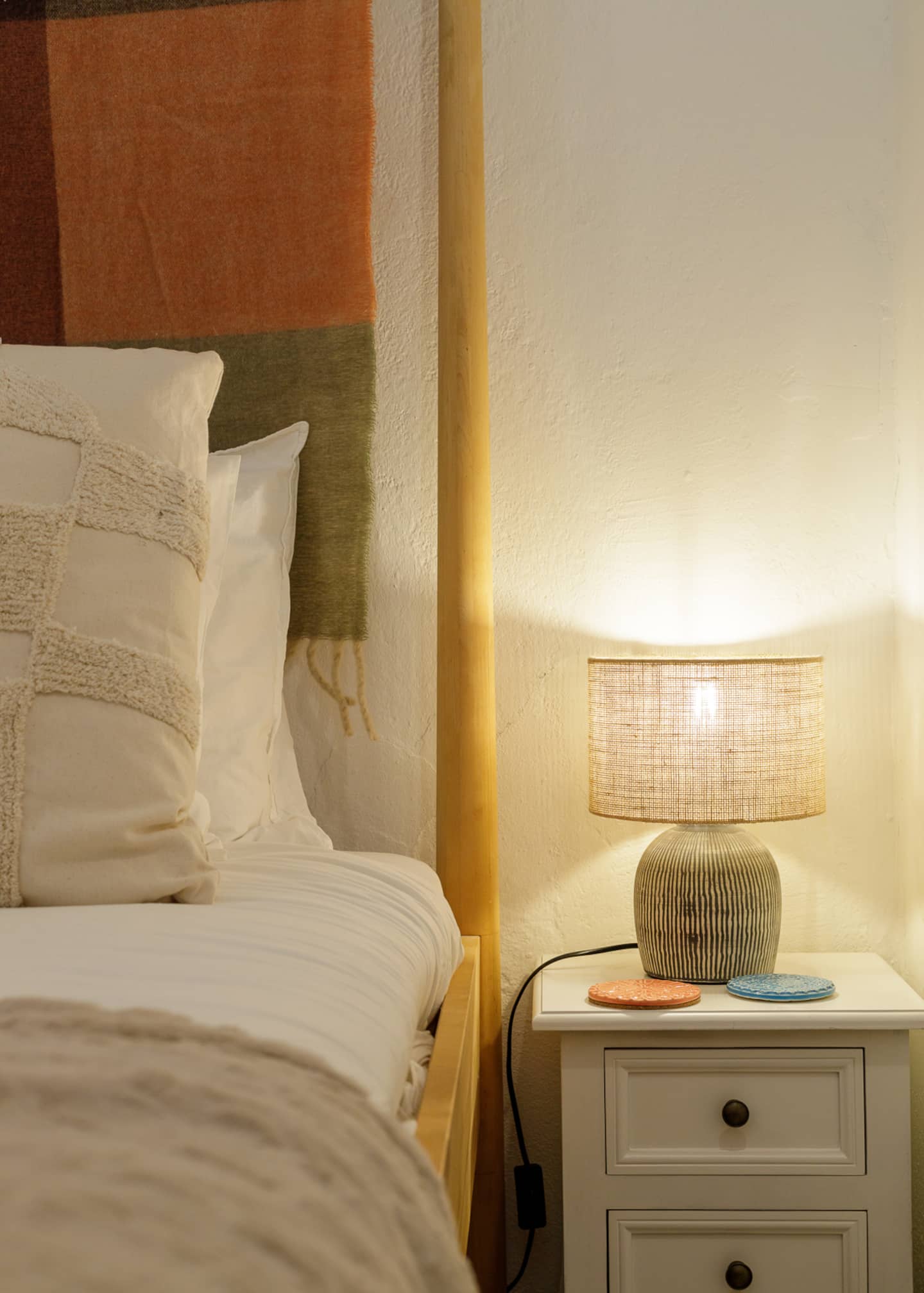 Beside the bed, a white bedside cabinet with a striped ceramic lamp lit under a linen shade, an orange and a blue coaster, a textured cream cushion and the striped blanket on the headboard.