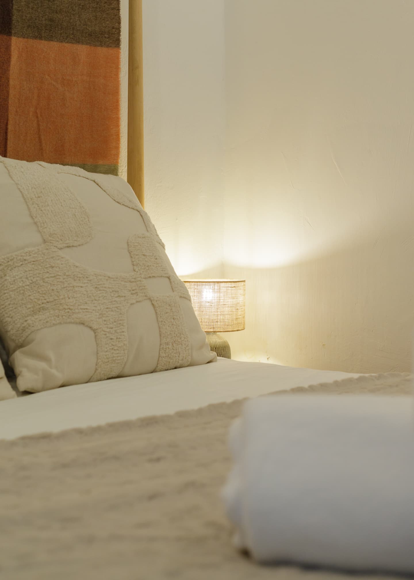 A cream cushion with a tufted pattern against the headboard blanket, a small bedside lamp glowing warm on the white plaster wall, and a folded white towel on the bed.