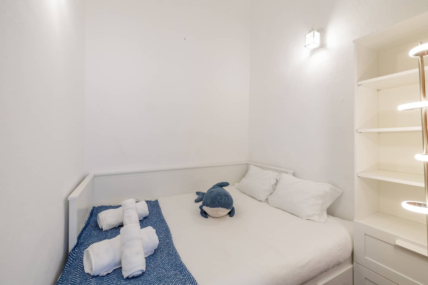 A small white bedroom: a bed with white pillows, a blue and white patterned throw, rolled white towels and a plush blue shark, a lantern wall light above, and white shelving on the right.