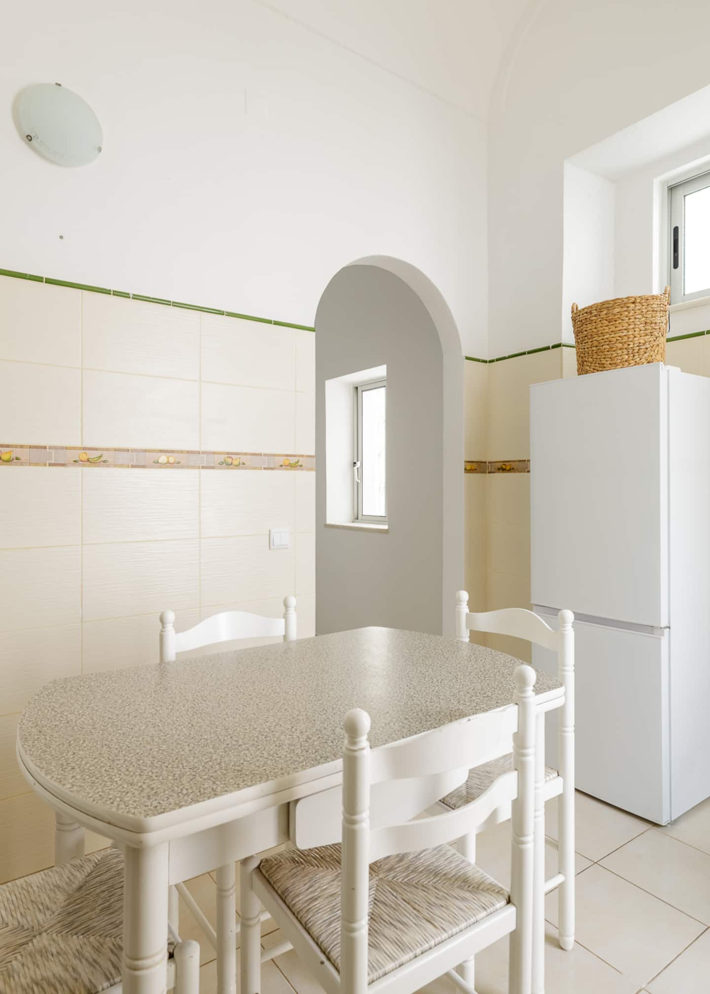A white dining table with a speckled top and rush-seated white chairs, an arched opening in the white wall with a small window beyond, a tall white fridge-freezer with a wicker basket on top, cream tiles with a green trim.