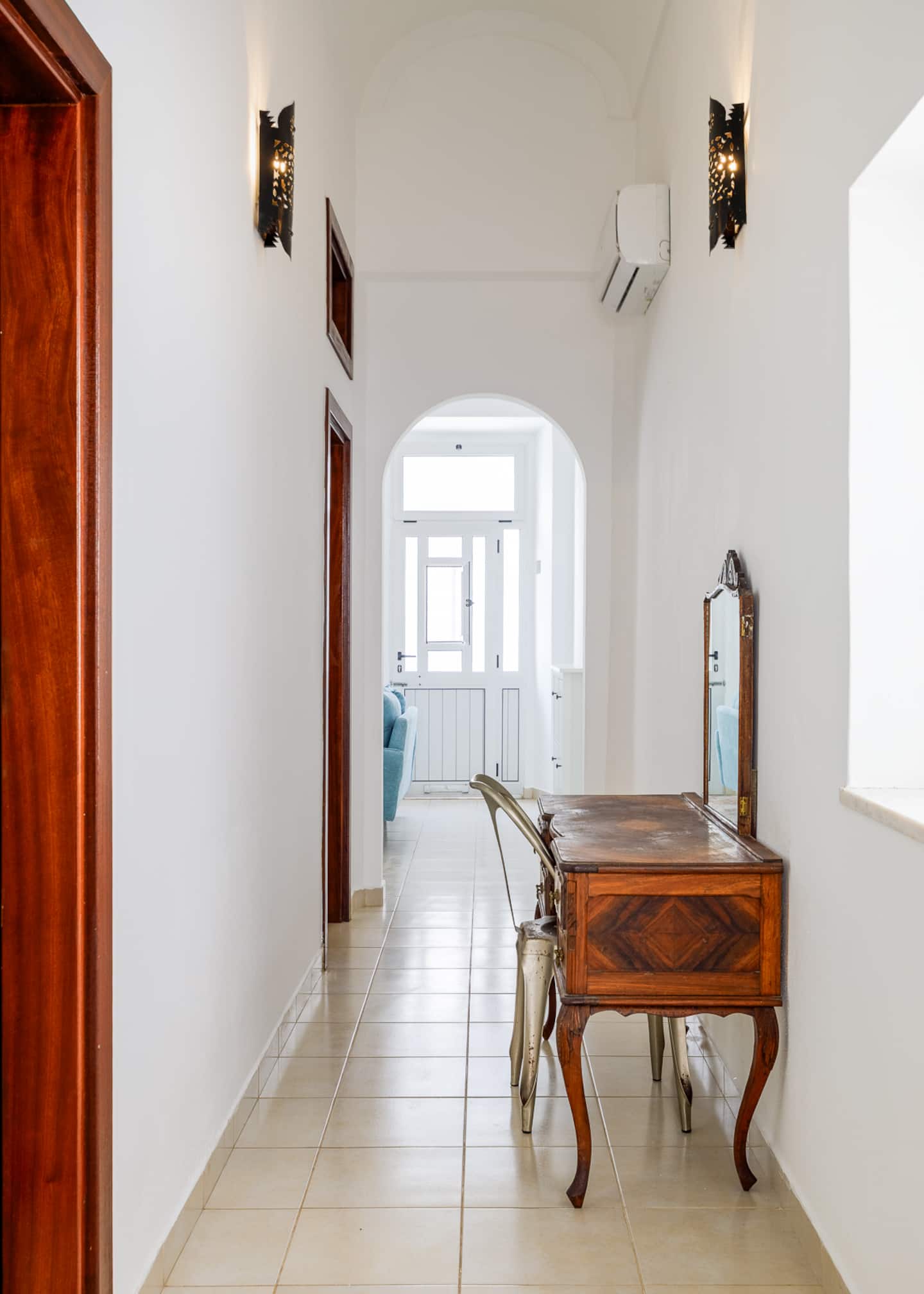 A long white hallway with a beige tiled floor and a vaulted ceiling, two pierced-metal wall lamps, doors of dark red timber, an antique wooden dressing table with a metal chair and a mirror, and an arched opening at the end onto the living room's blue sofa.