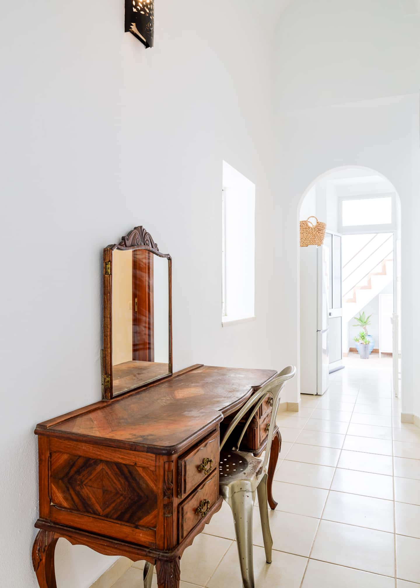 An antique wooden dressing table with a hinged mirror and a metal café chair against a white wall; beyond an arch, the kitchen's fridge with a wicker basket on top and, through a glazed door, the exterior stair.