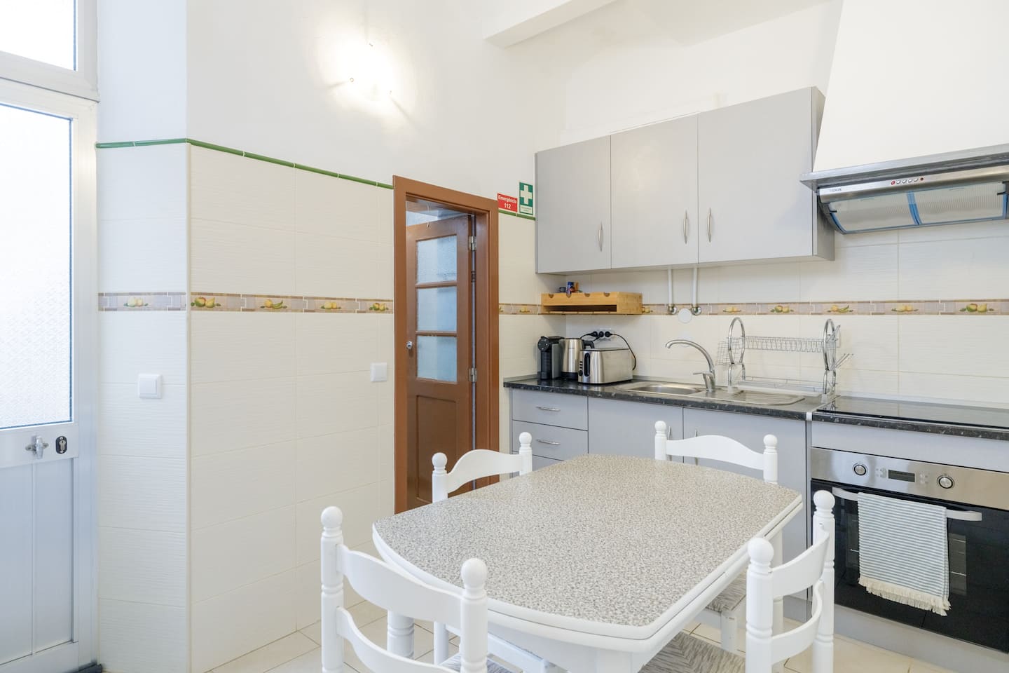 A kitchen with cream tiled walls and a fruit-patterned tile border: grey cabinets, a sink and hob under a stainless hood, a built-in oven, a white table with four white chairs, and a glazed door in dark red timber.