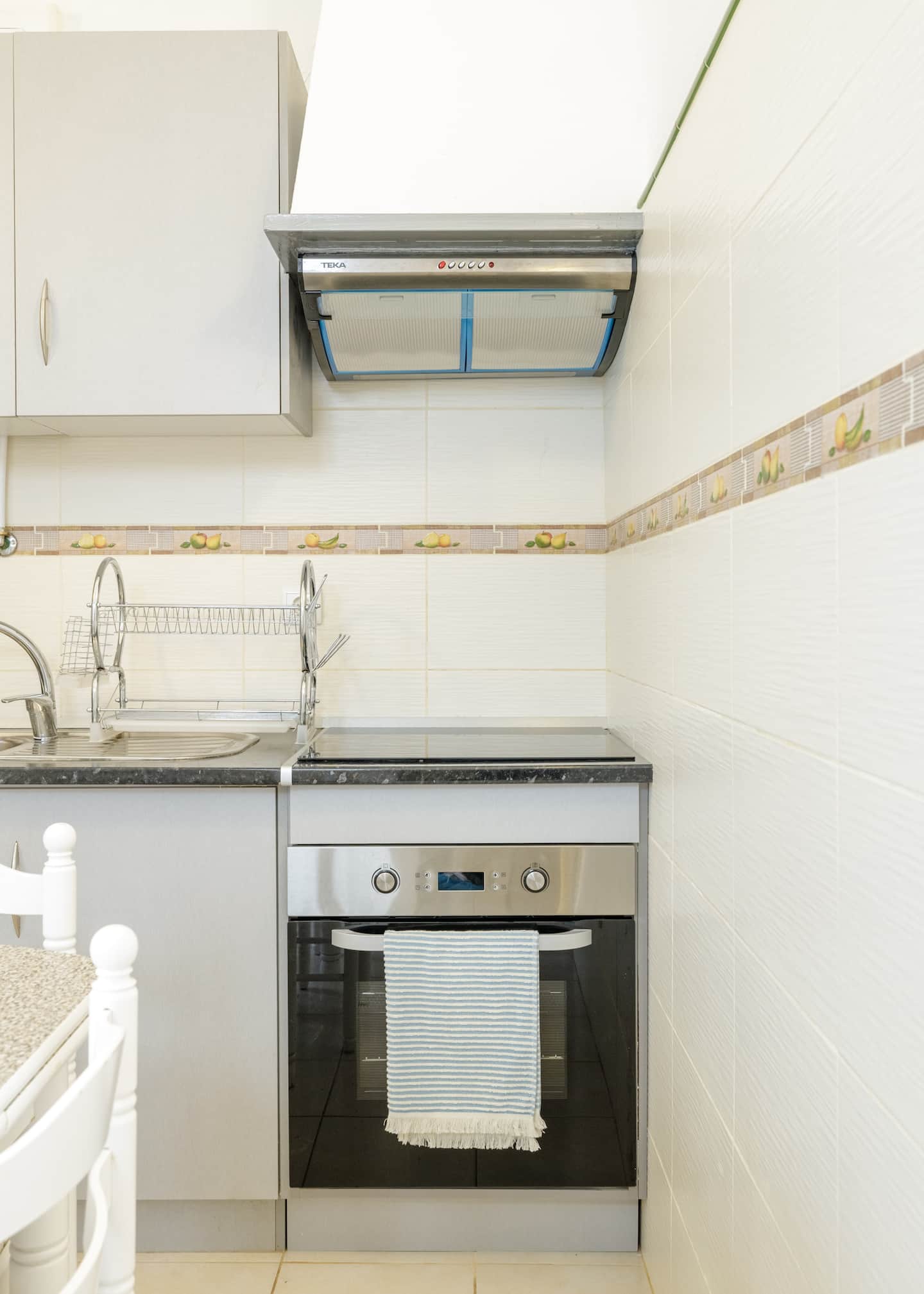 A ceramic hob and a stainless steel oven set into grey cabinets under an extractor hood, cream tiles with a border of lemons and fruit, a dish rack by the sink, and a striped tea towel on the oven handle.