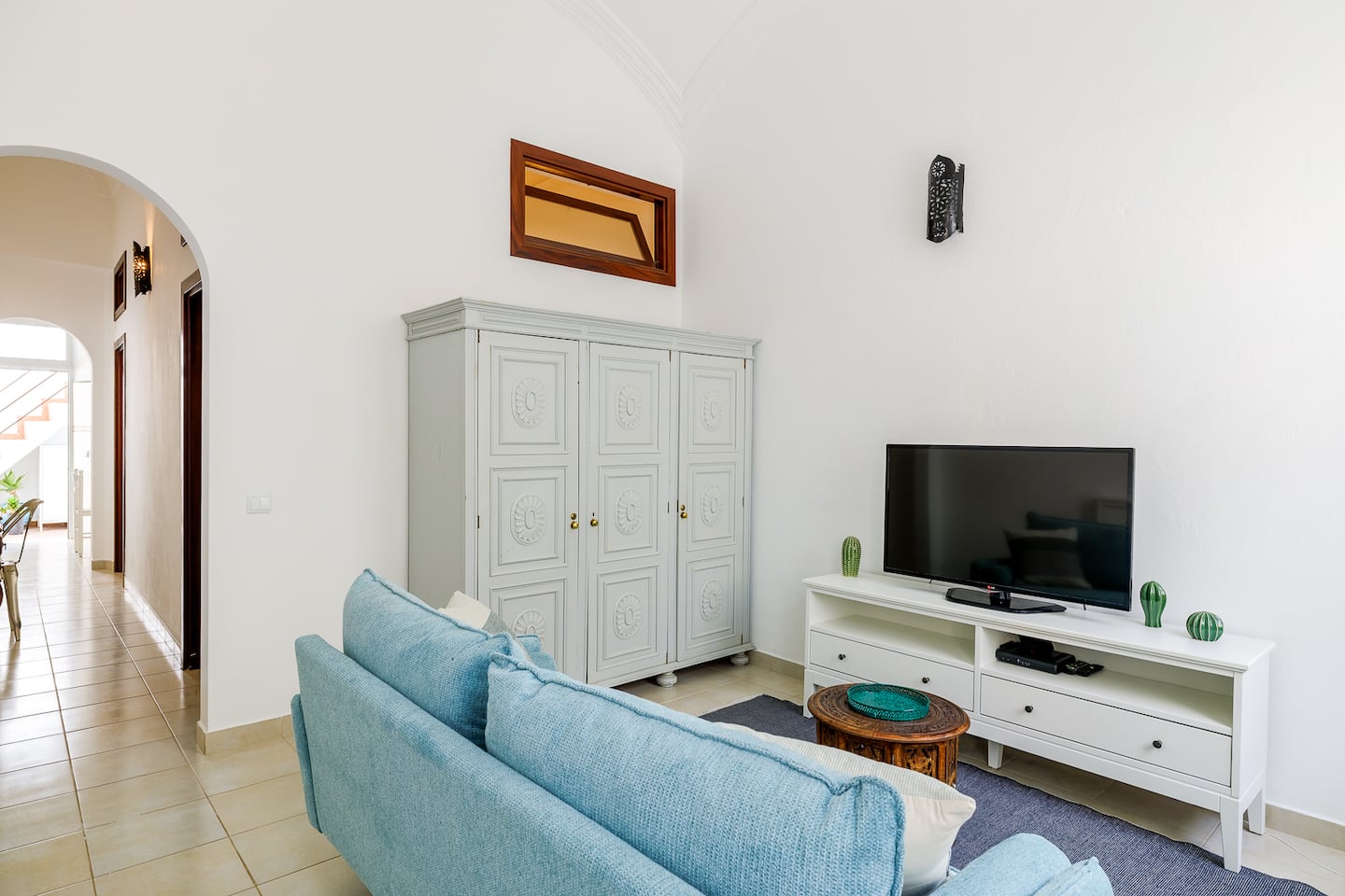 A living room with white walls and a beige tiled floor: a sky-blue sofa on a grey rug faces a television on a white cabinet; behind it a pale grey wardrobe with carved panels, and an arched doorway on the left opens to a hallway.