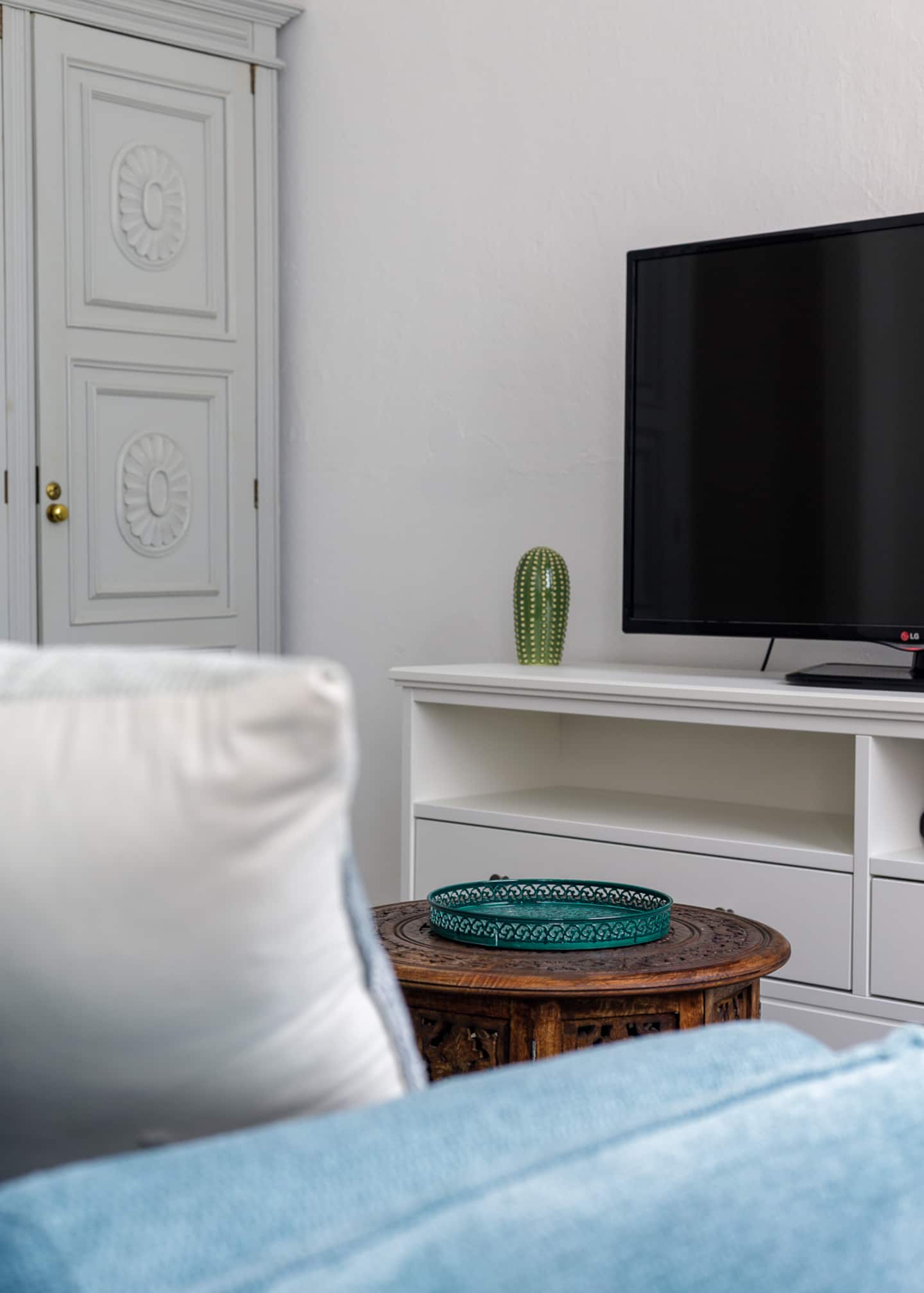 Past a white cushion on the blue sofa: a round carved wooden side table with a teal tray, a pale grey wardrobe door with carved rosettes, a cactus ornament and a television on a white unit.