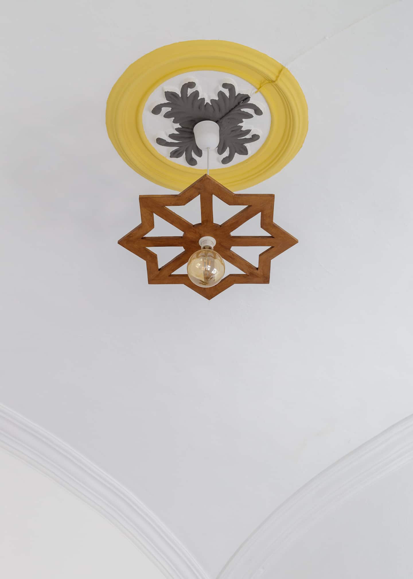 Looking up at a white vaulted ceiling: a yellow plaster ceiling rose with grey leaf mouldings, and a wooden eight-point star light fitting with a bare filament bulb.