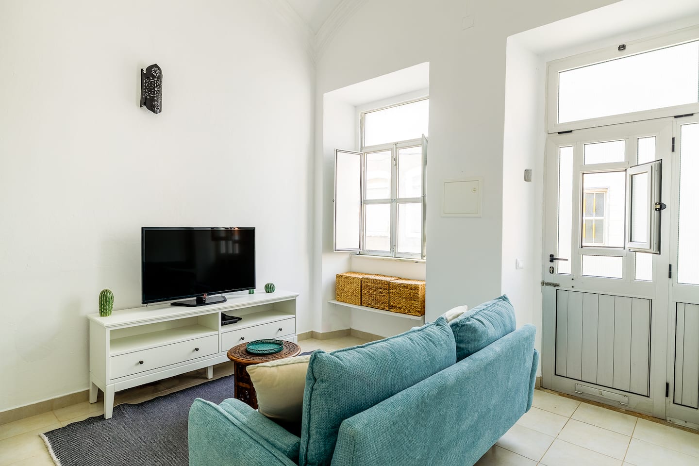 The living room towards the street: a deep-set window with its shutters open and wicker baskets on the sill, a half-glazed white front door, the blue sofa and a television on a white cabinet.