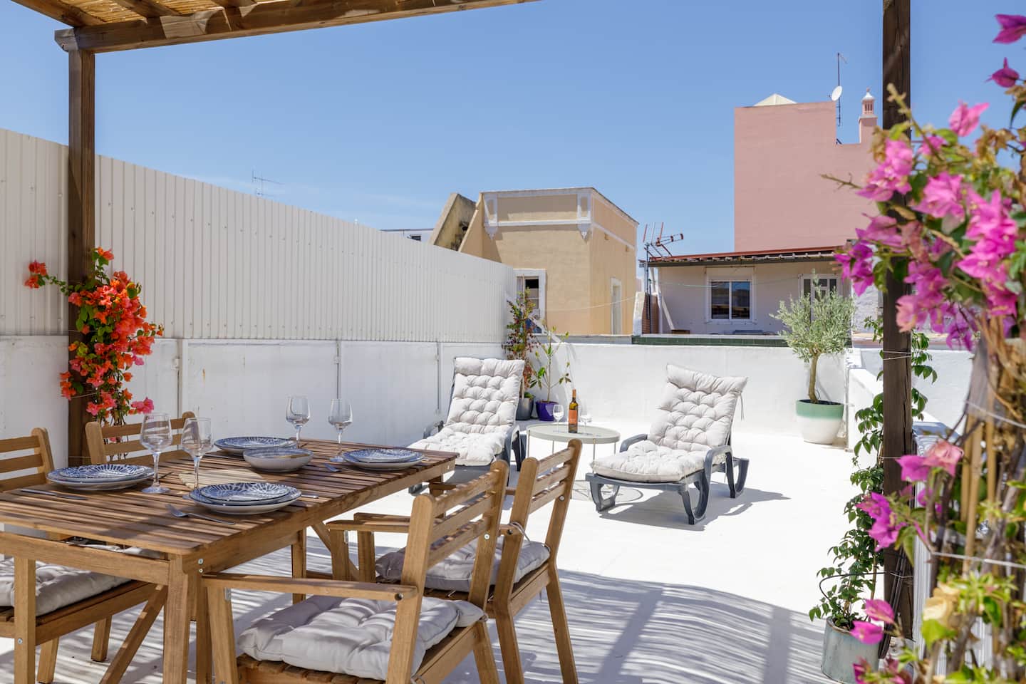 A whitewashed roof terrace under a cane-and-timber pergola: a wooden table set for four with blue-patterned plates and glasses, two cushioned loungers by a round table with a bottle of wine, a small olive tree in a pot, magenta bougainvillea on the right and orange bougainvillea on the left, rooftops and a blue sky beyond.