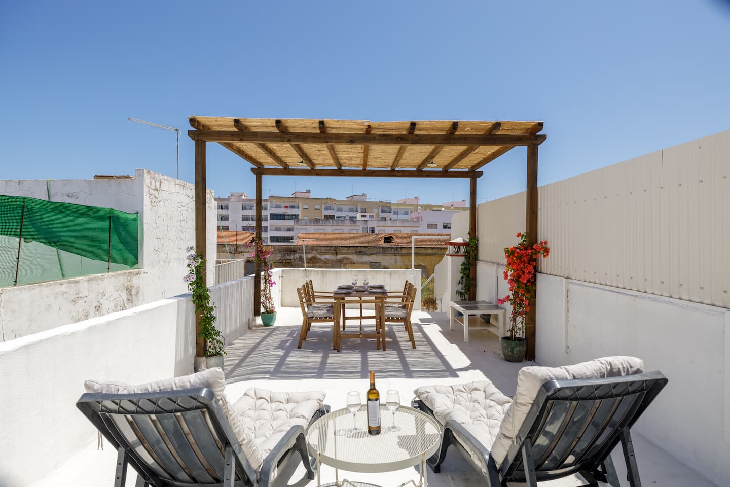 The roof terrace seen from the loungers: two cushioned loungers either side of a round glass table with a bottle of wine and two glasses, the timber pergola over a wooden dining table and chairs, red bougainvillea by the pergola posts, white walls and the town's rooftops under a deep blue sky.