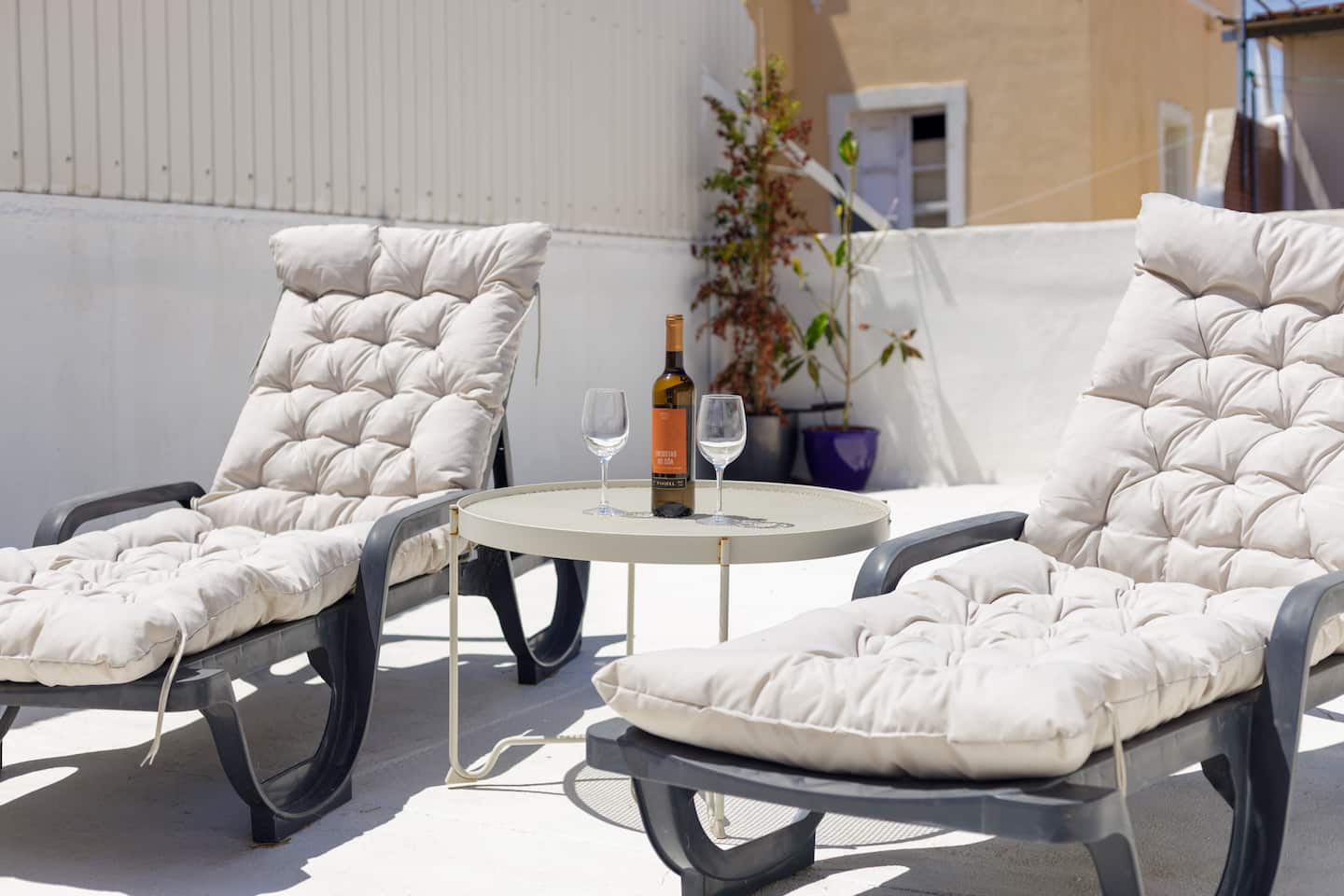 Two cushioned loungers with a small round table between them holding a bottle of white wine and two glasses, on a white terrace floor, with a potted shrub, a purple pot and the neighbouring yellow house behind.