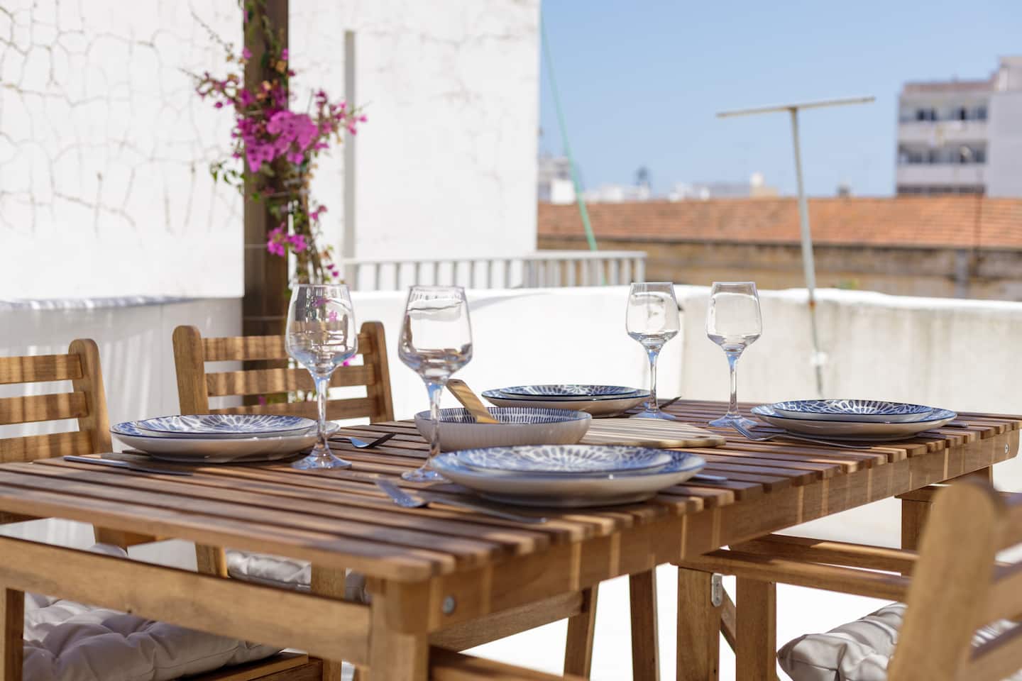 A slatted wooden table laid with blue-patterned plates, bowls and four wine glasses, magenta bougainvillea over the white wall behind, and the red-tiled rooftops of Olhão beyond.