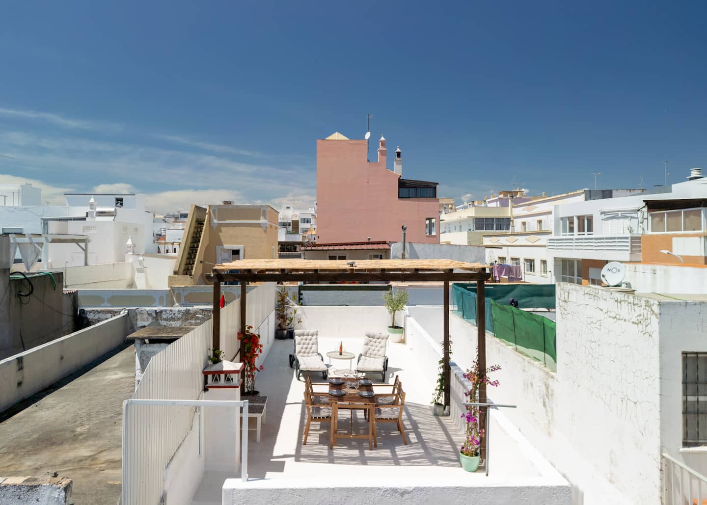 The roof terrace seen from above: the timber pergola over the dining table, two loungers and a round table on the white floor, walls around it, and the pink and white buildings of Olhão under a blue sky.